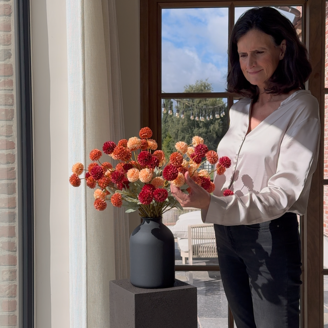 A woman arranges a bouquet of red and orange pom-pom flowers in a black vase near a window.