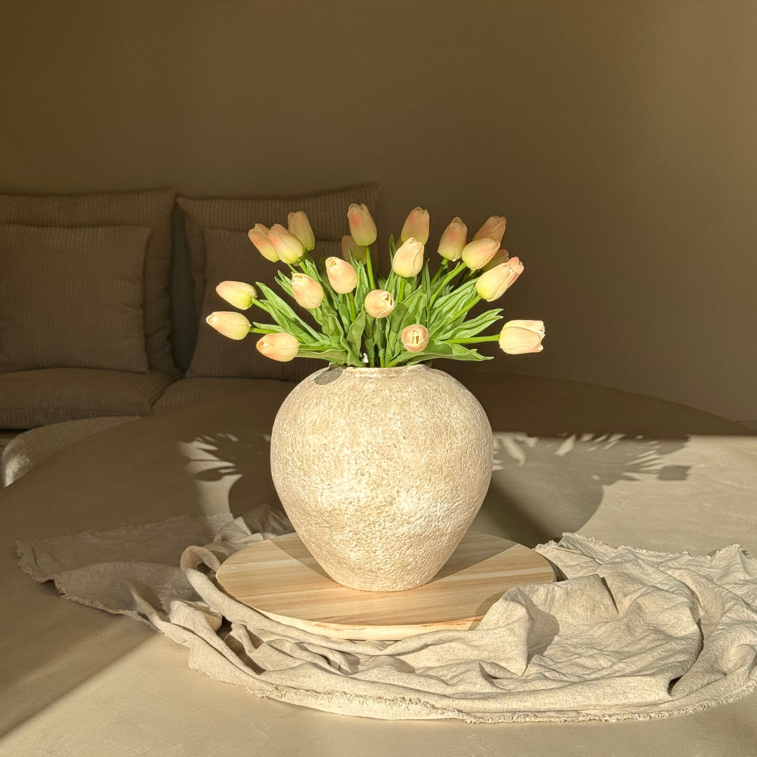 A bouquet of pink tulips in a round, textured beige vase sitting on a bed in the sunlight.
