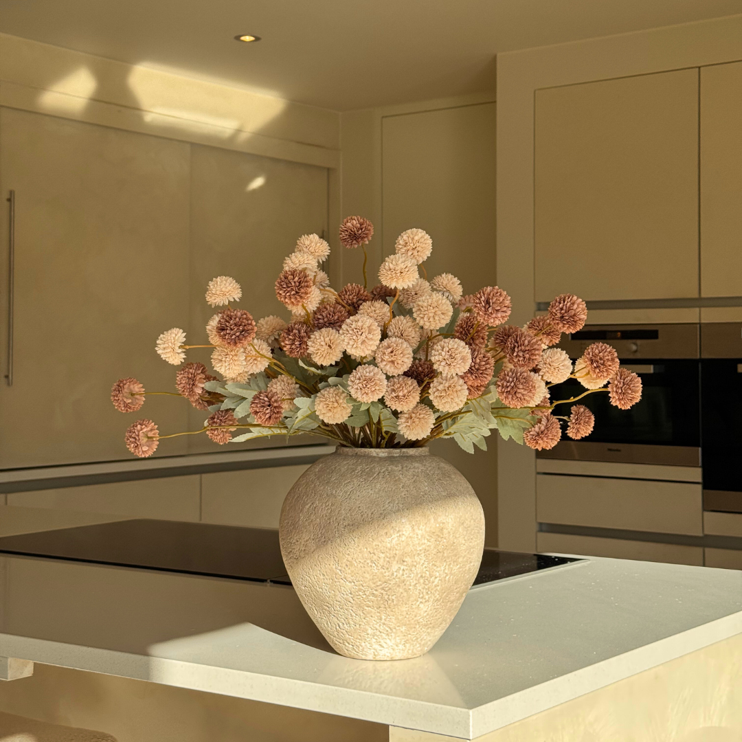 A textured beige vase with pink and cream pom-pom flowers sits on a sunlit kitchen island.