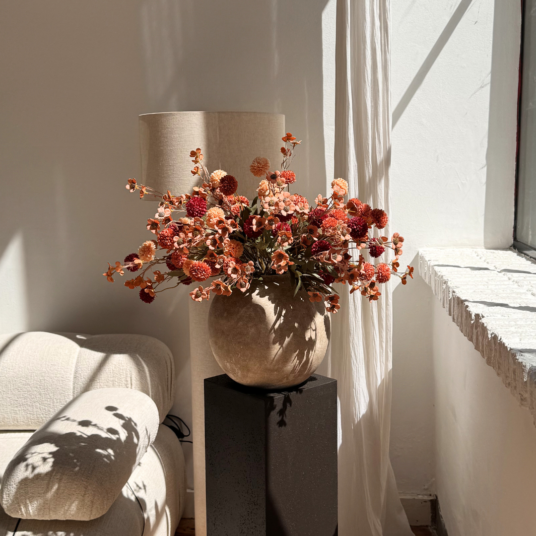 A round vase with reddish-orange flowers sits on a dark pedestal in a room lit by sunlight.