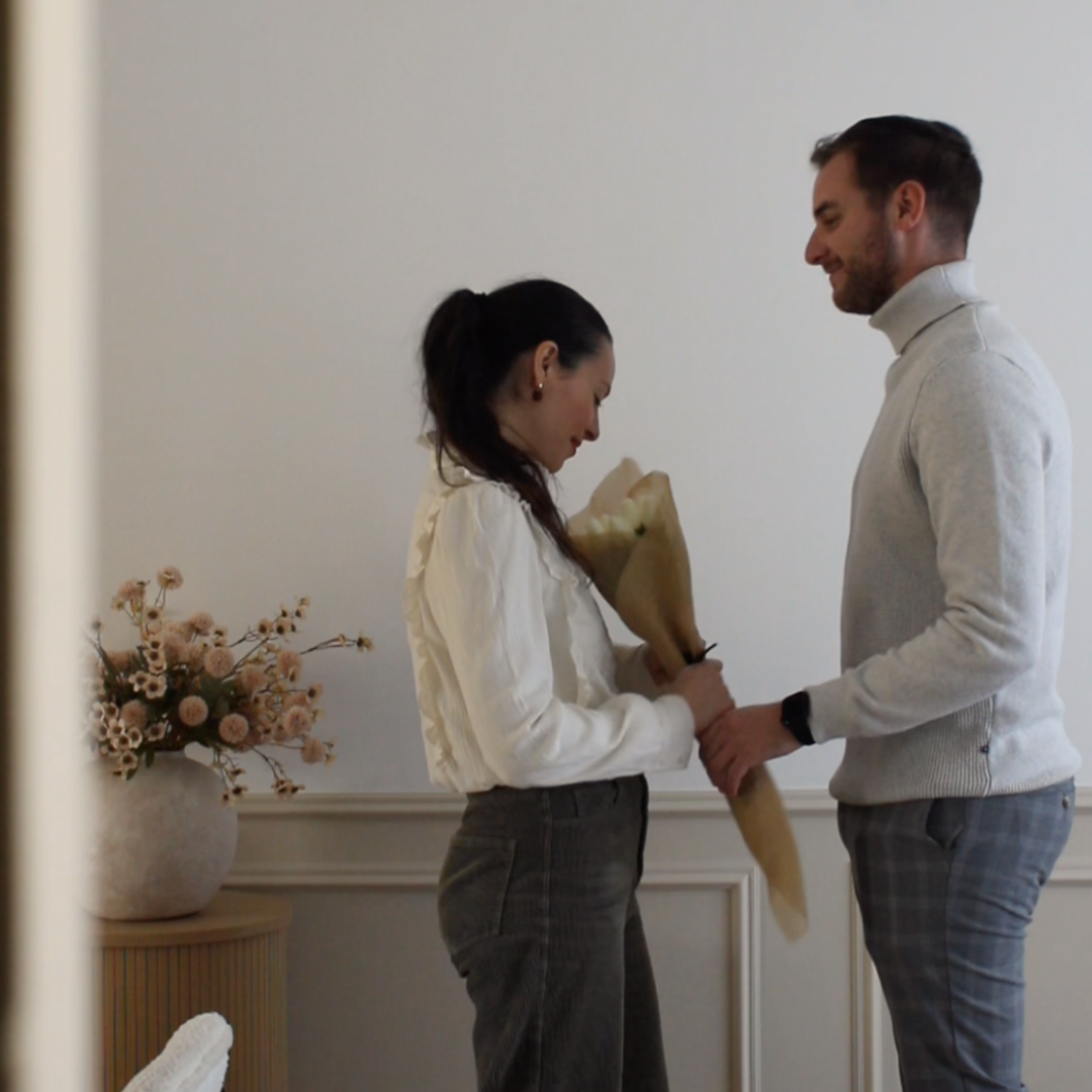 A man in a light grey turtleneck sweater gives a bouquet of flowers to a woman.