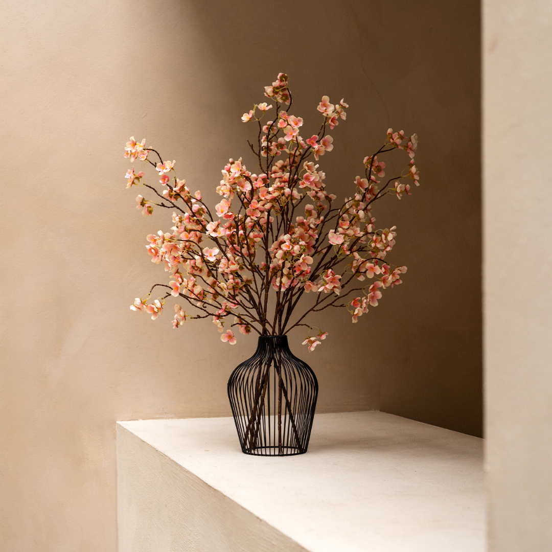 Wire vase with pink cherry blossoms on a white ledge.