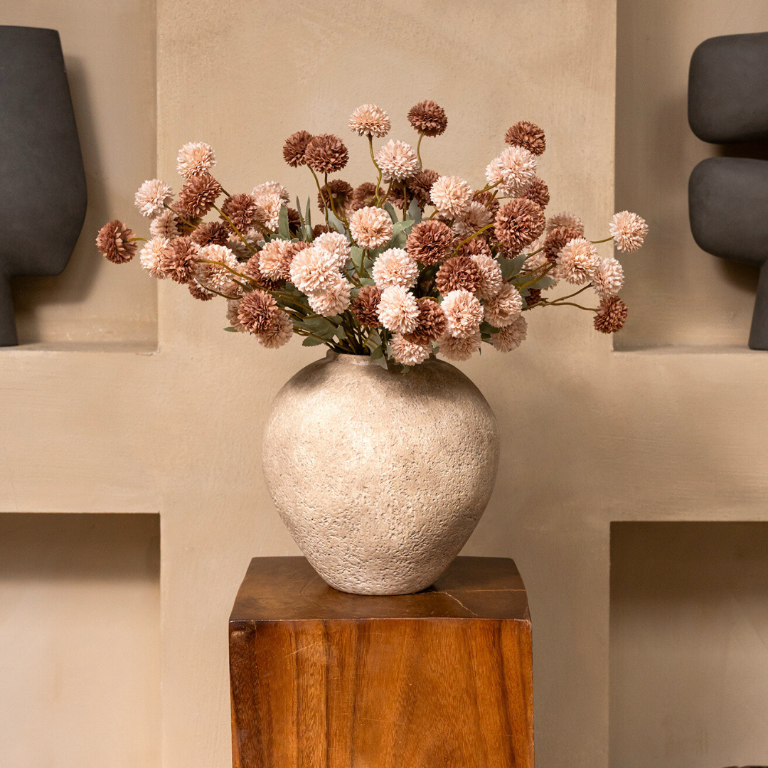 Pink and brown pom-pom flowers in a textured, round vase resting on a wooden block.