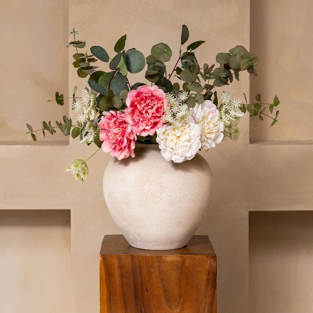 A textured vase with pink and white flowers and green eucalyptus leaves on a wooden stand.