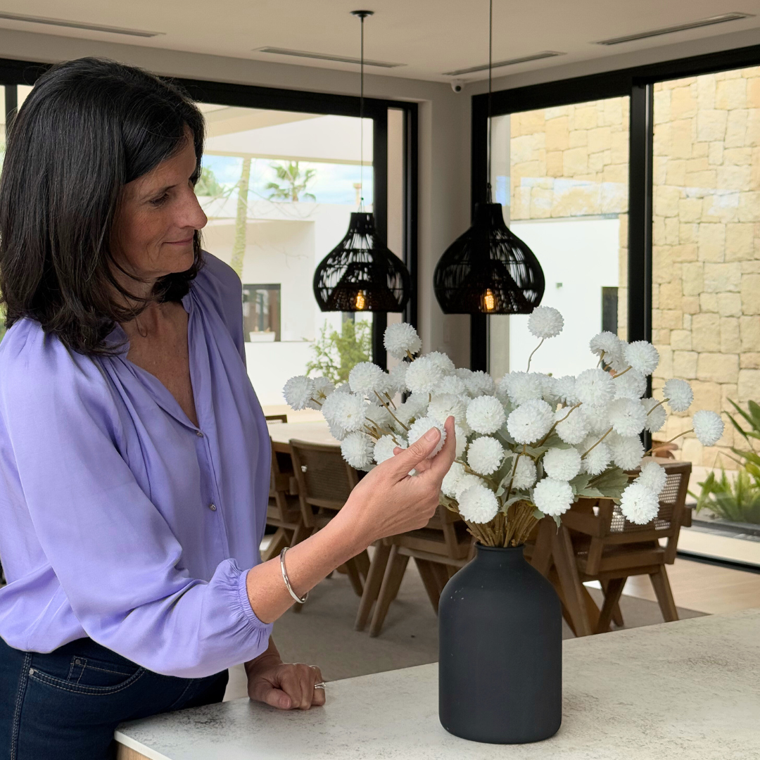 Woman in purple shirt arranging white flowers in a black vase indoors.