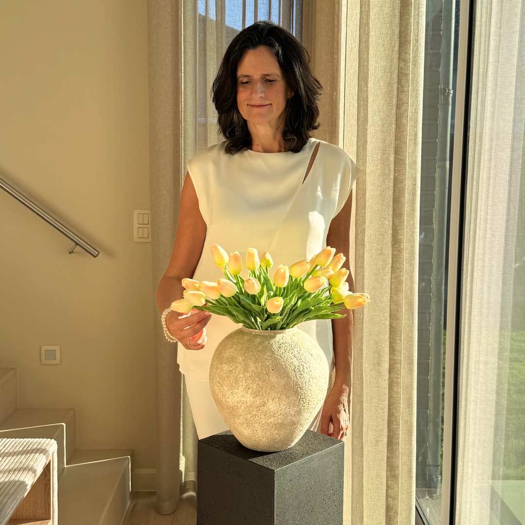 Woman in white dress arranging yellow tulips in a round vase indoors.