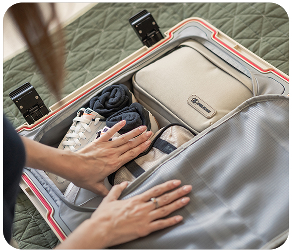 Hands packing a suitcase with shoes and a beige pouch on a green surface.