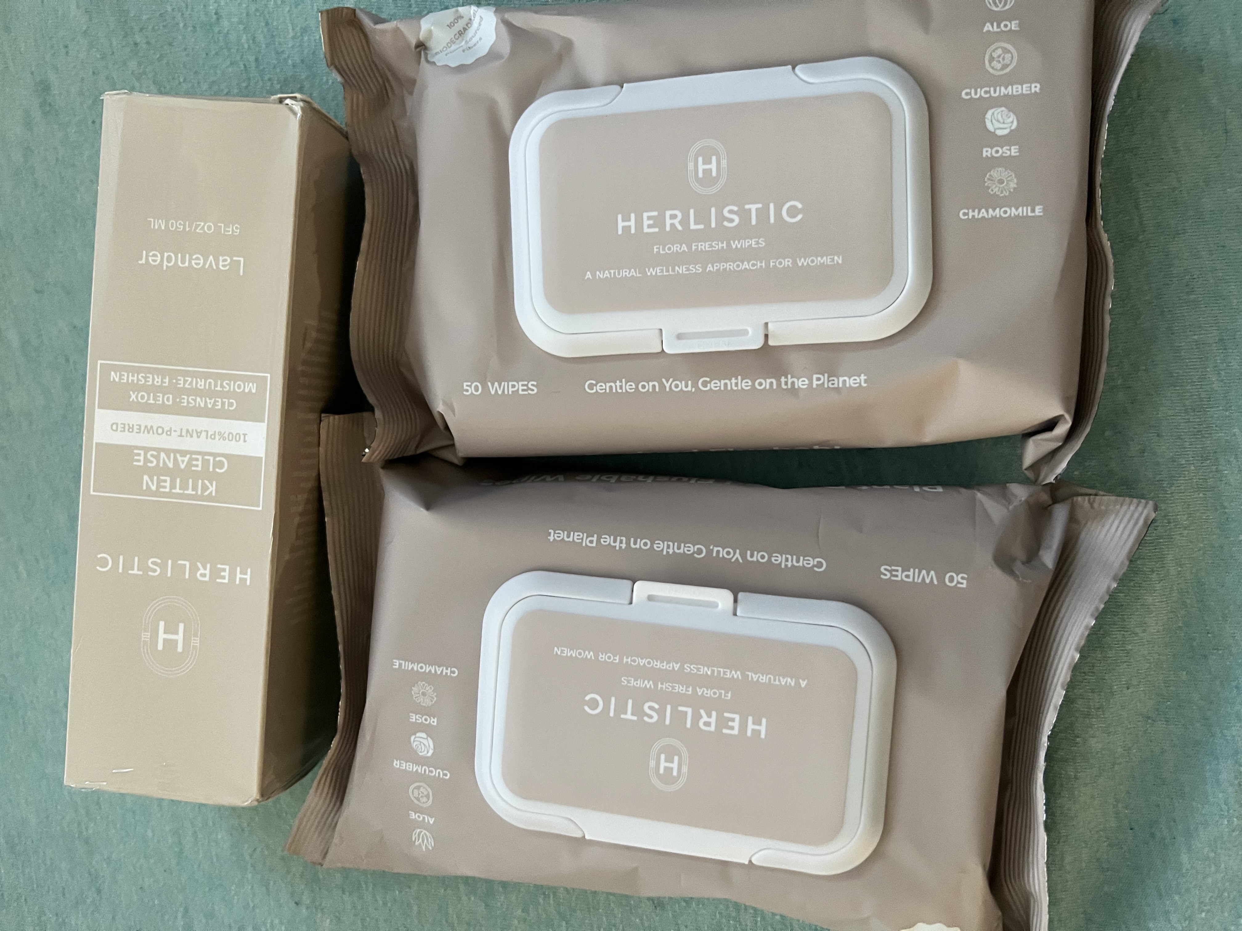 Two packs of Herlistic wipes and a box of Kitten Cleanse on a teal surface.