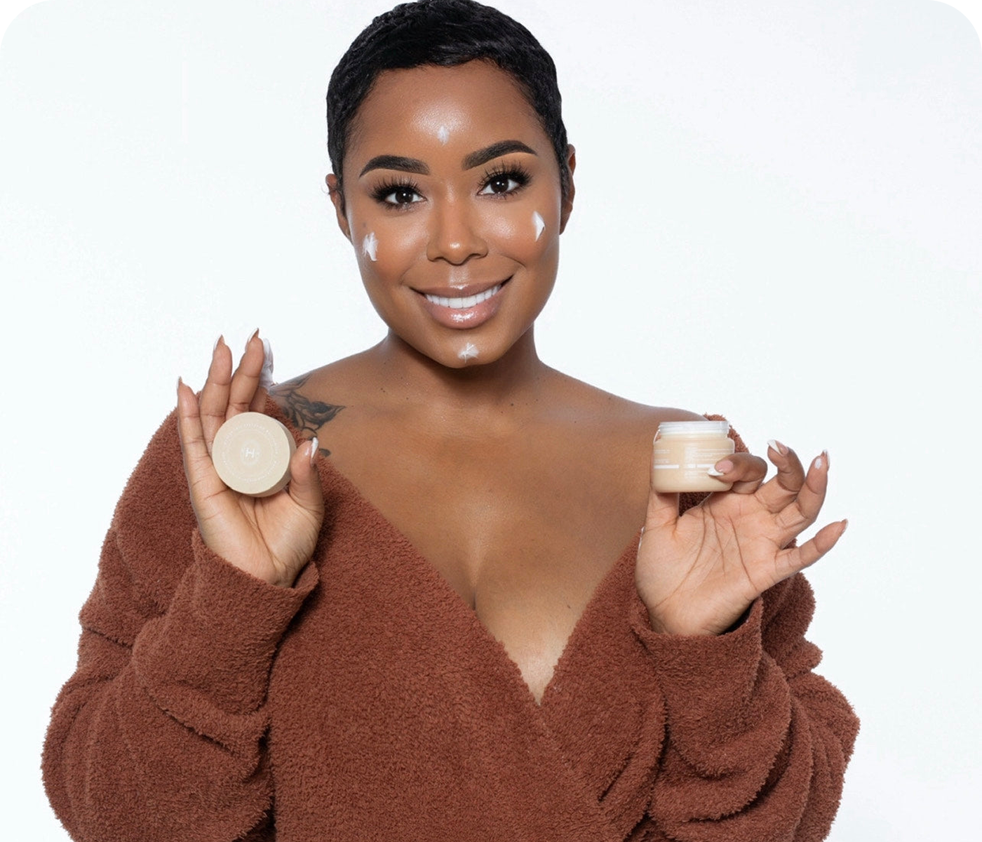 Person smiling, holding skincare products, with cream applied on face.