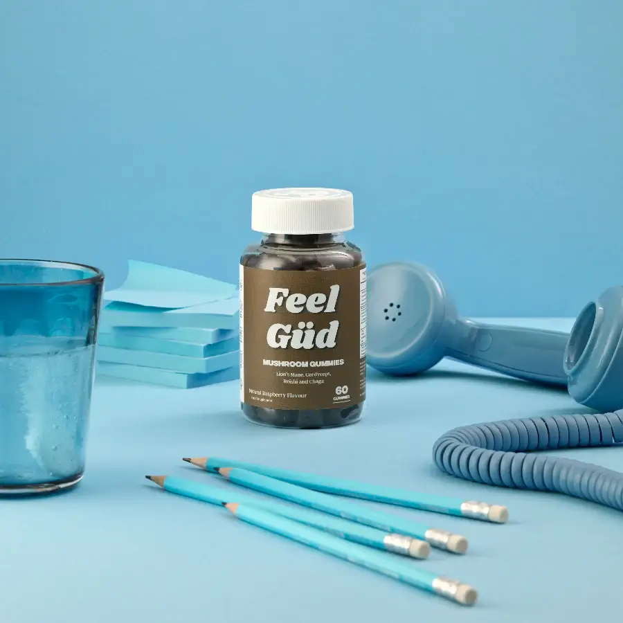 Bottle of 'Feel Gǖd' mushroom gummies on a blue desk with pencils and a phone.