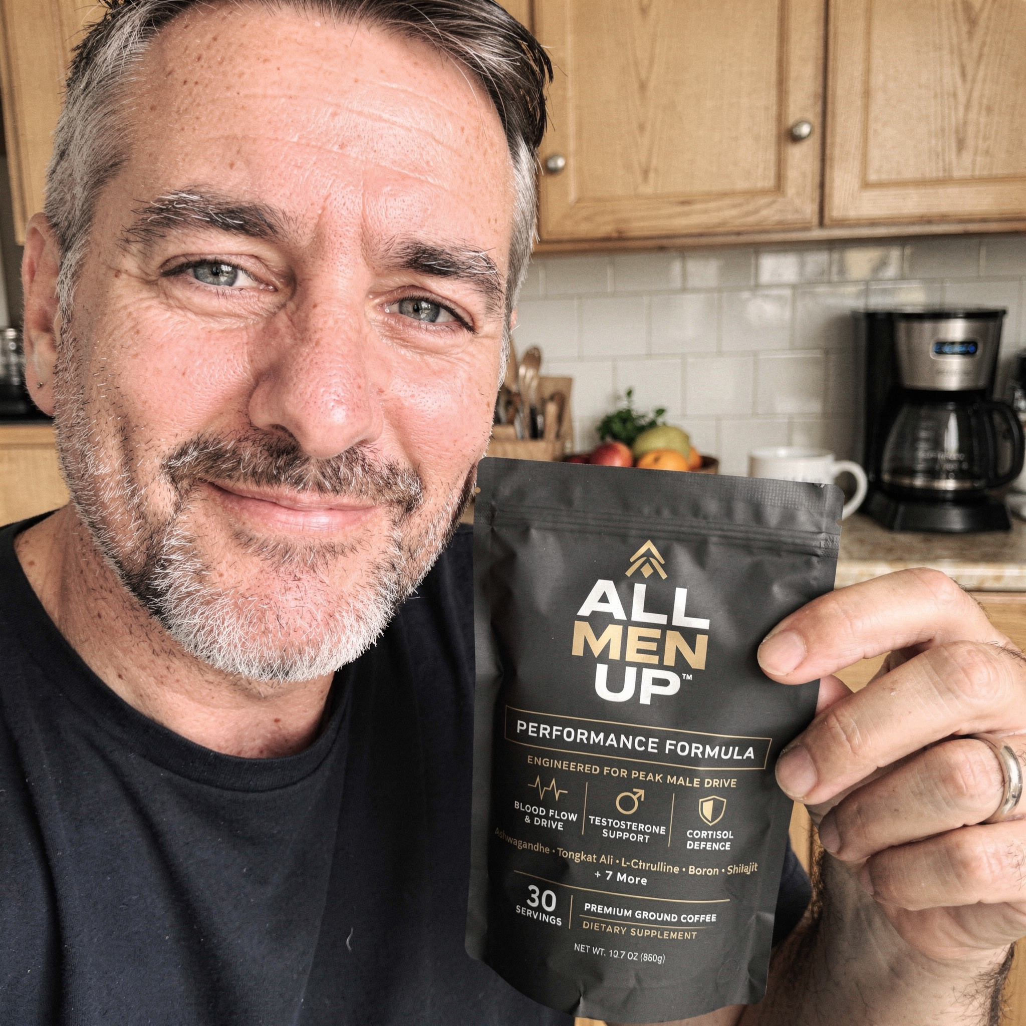 A middle-aged man with a graying beard holds up a black pouch of All Men Up supplement.