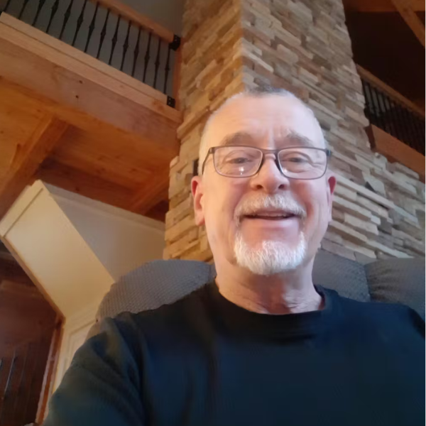 A smiling man with glasses and a goatee sitting indoors in front of a stone fireplace.