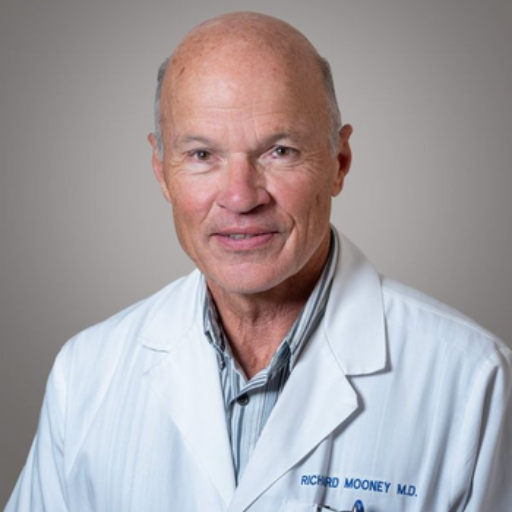A headshot of an older, bald man with brown eyes, wearing a white lab coat and smiling.
