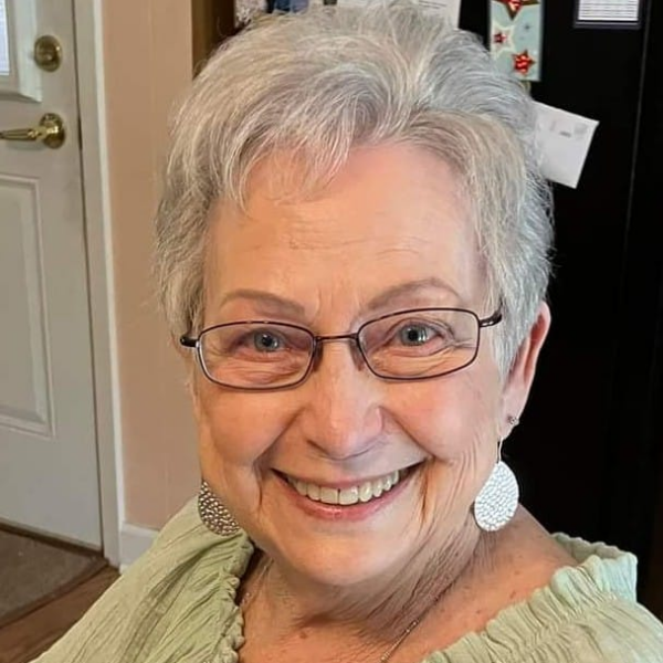 A smiling older woman with short gray hair and glasses, wearing a light green shirt and large earrings.