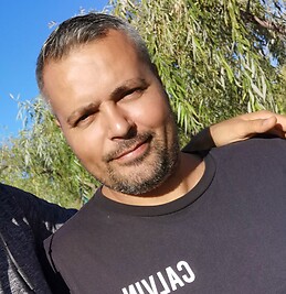 A close-up of a man with salt-and-pepper hair and a beard, smiling outdoors in a black t-shirt.