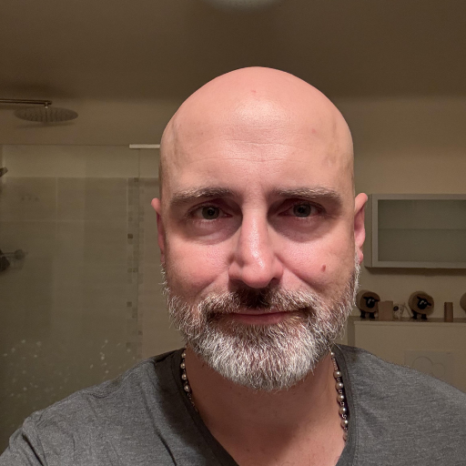 A close-up photo of a bald man with a salt and pepper beard, looking directly at the camera.