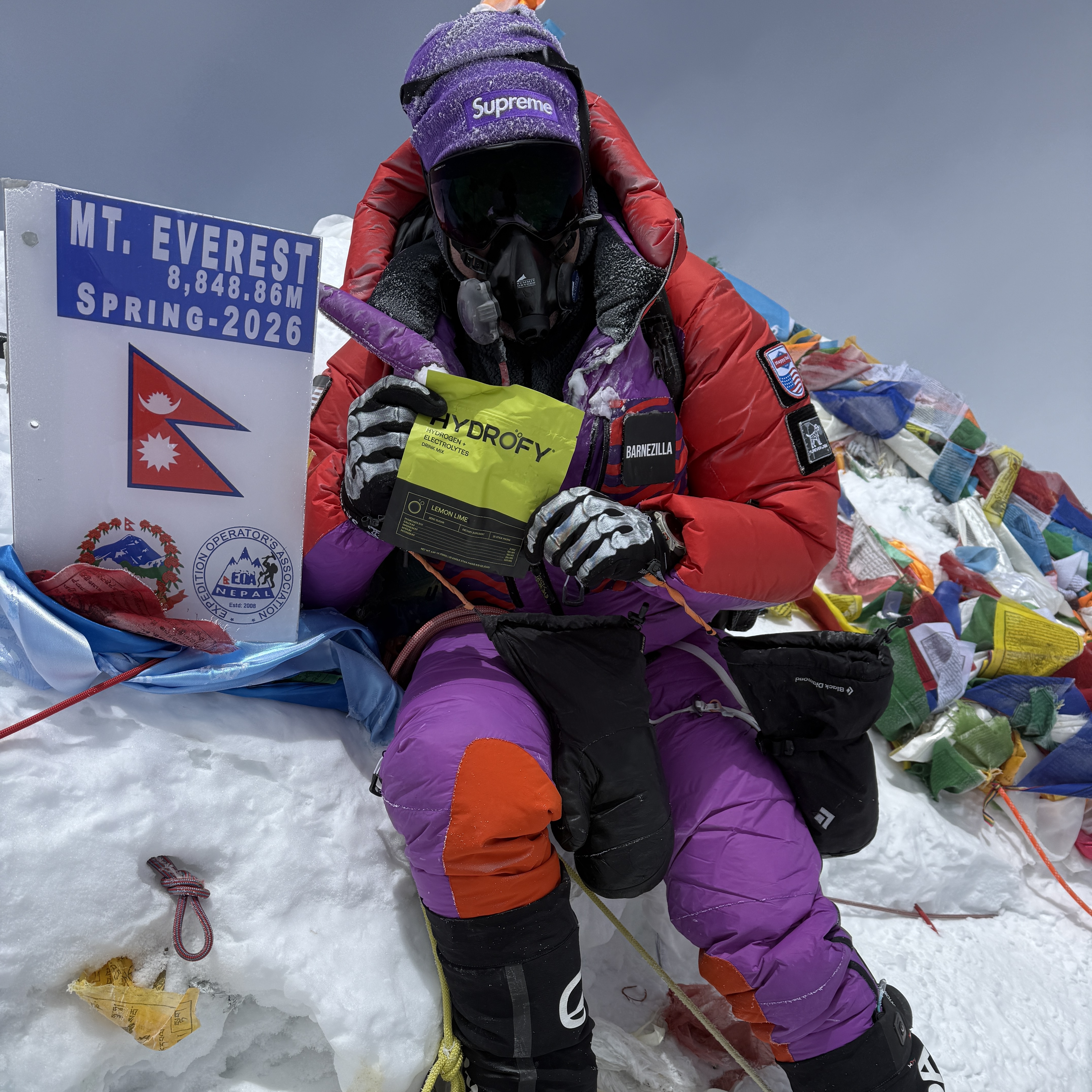 A climber in full gear with an oxygen mask sits on the snowy summit of Mt. Everest.