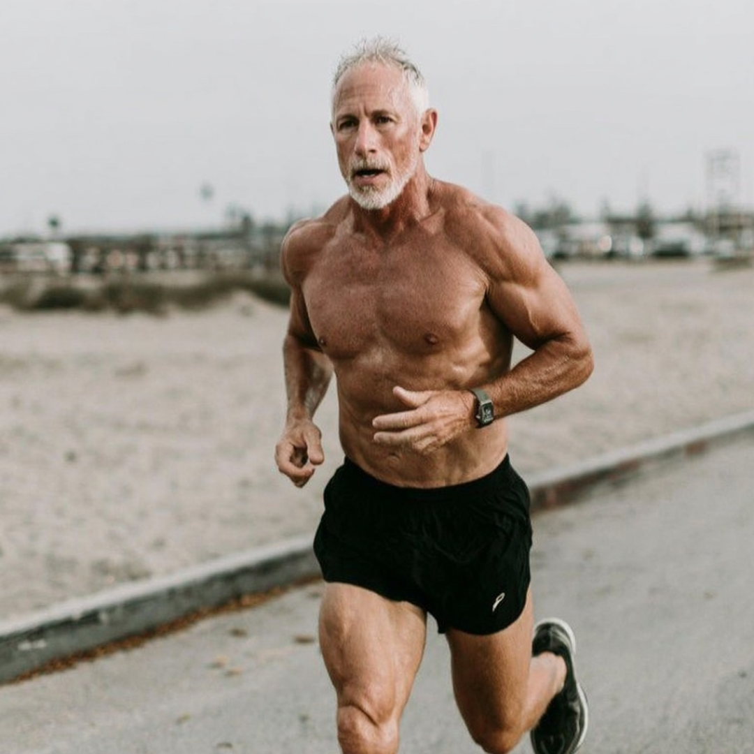 Shirtless man with gray hair running on a beachside path.