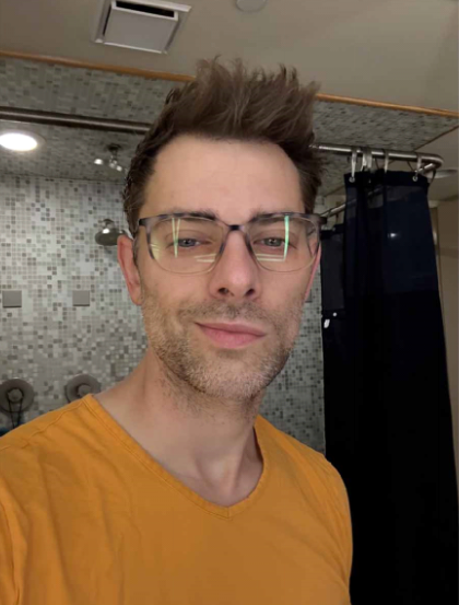 Person wearing glasses and an orange shirt in a bathroom with tiled walls.