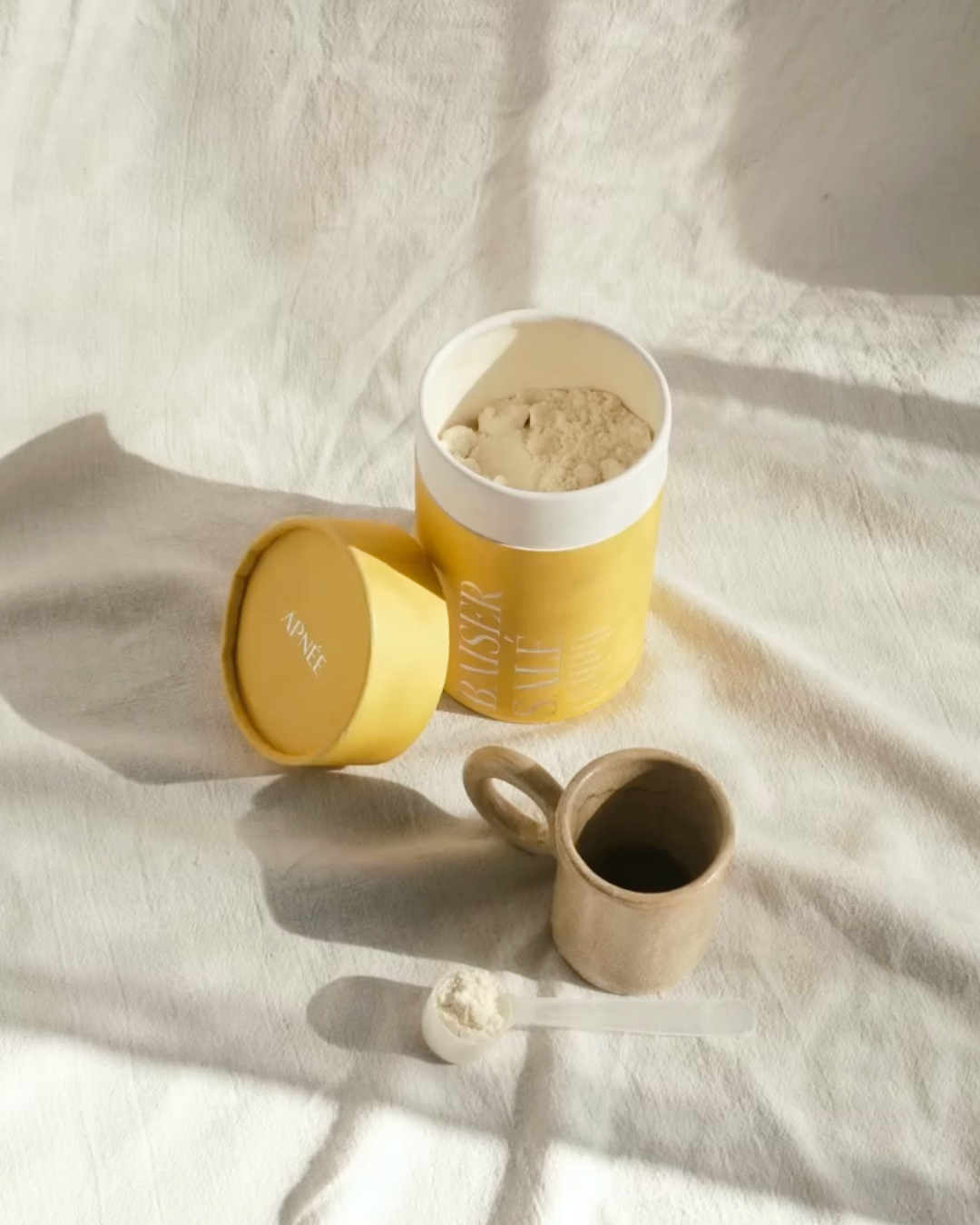 Yellow container with powder, ceramic mug, and scoop on a white cloth.