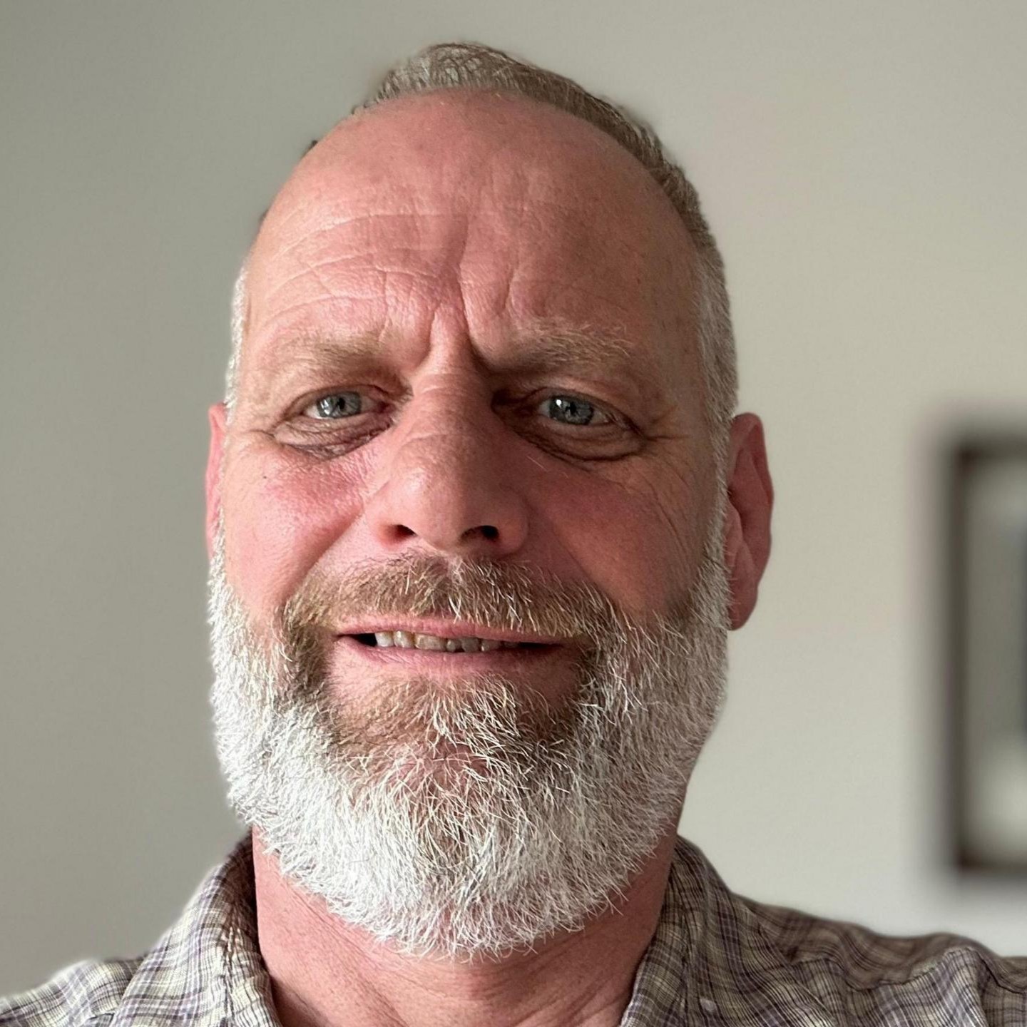A close-up photo of a middle-aged man with a gray beard and blue eyes, wearing a plaid shirt.