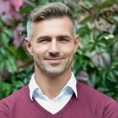 A man with salt-and-pepper hair and a maroon sweater smiles in front of leafy foliage.