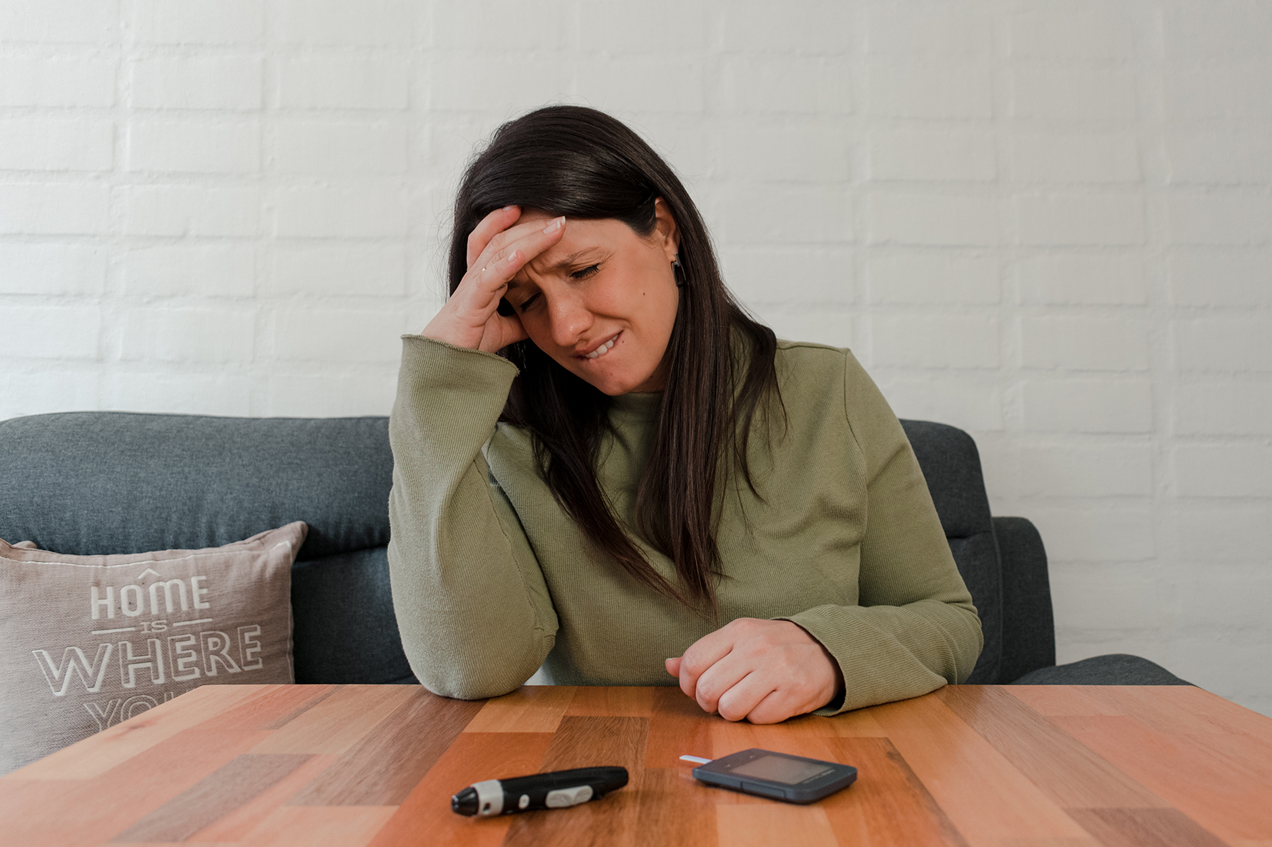 A distressed woman sits at a table with her hand on her head, a glucometer before her.