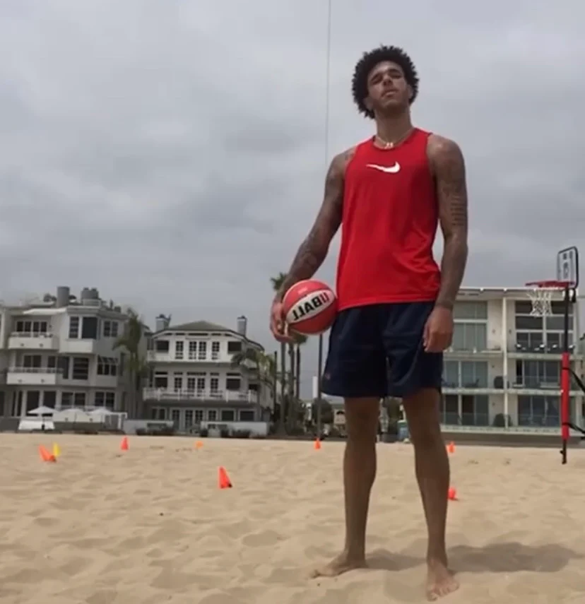 Person in red tank top holding a basketball on a sandy beach with buildings in background.