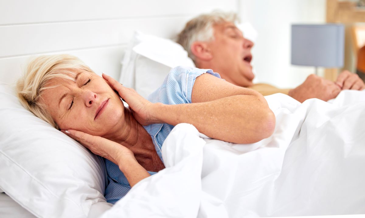 A woman lies in bed covering her ears while a man snores next to her.