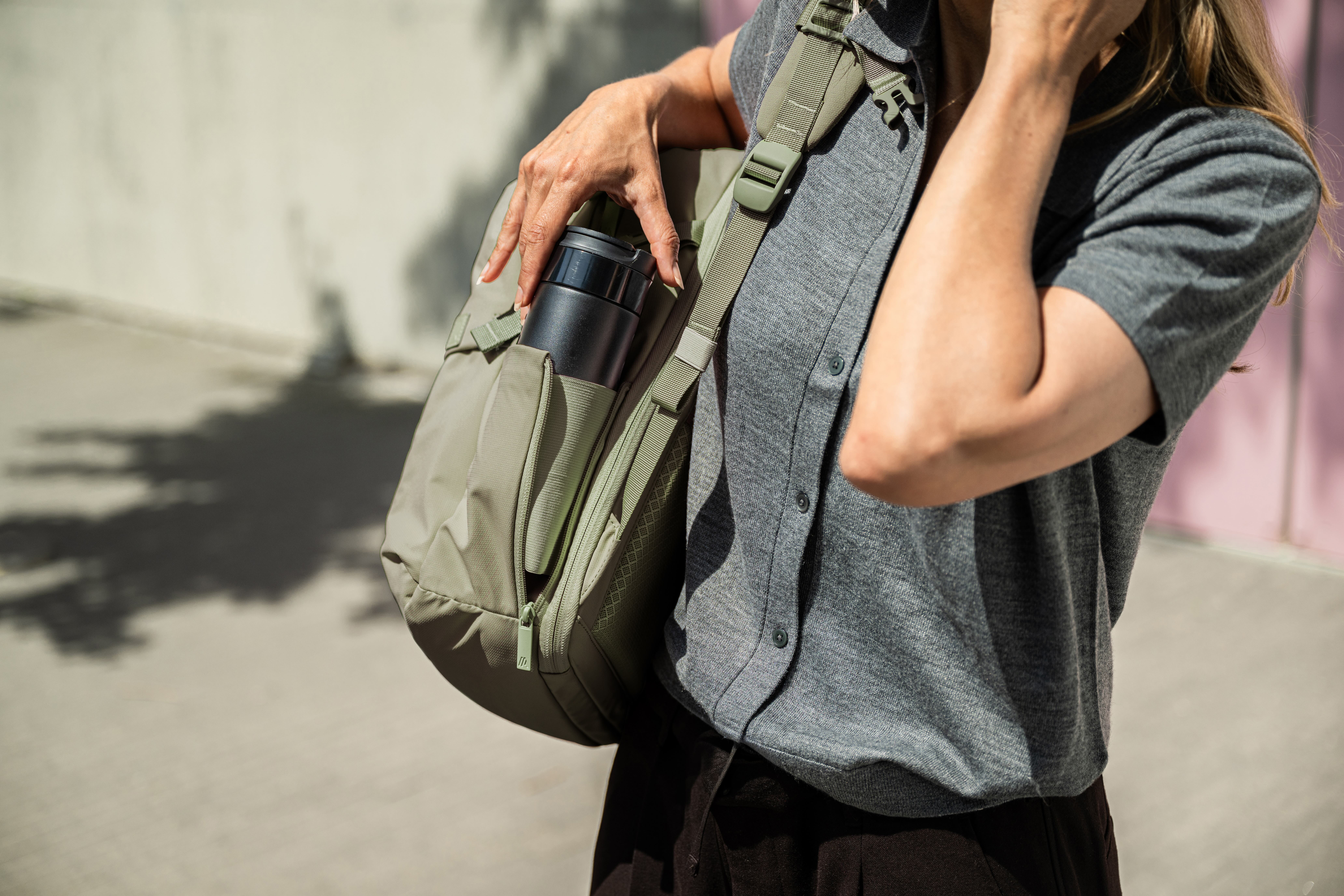 A person places a black travel mug into the side pocket of a light green backpack.
