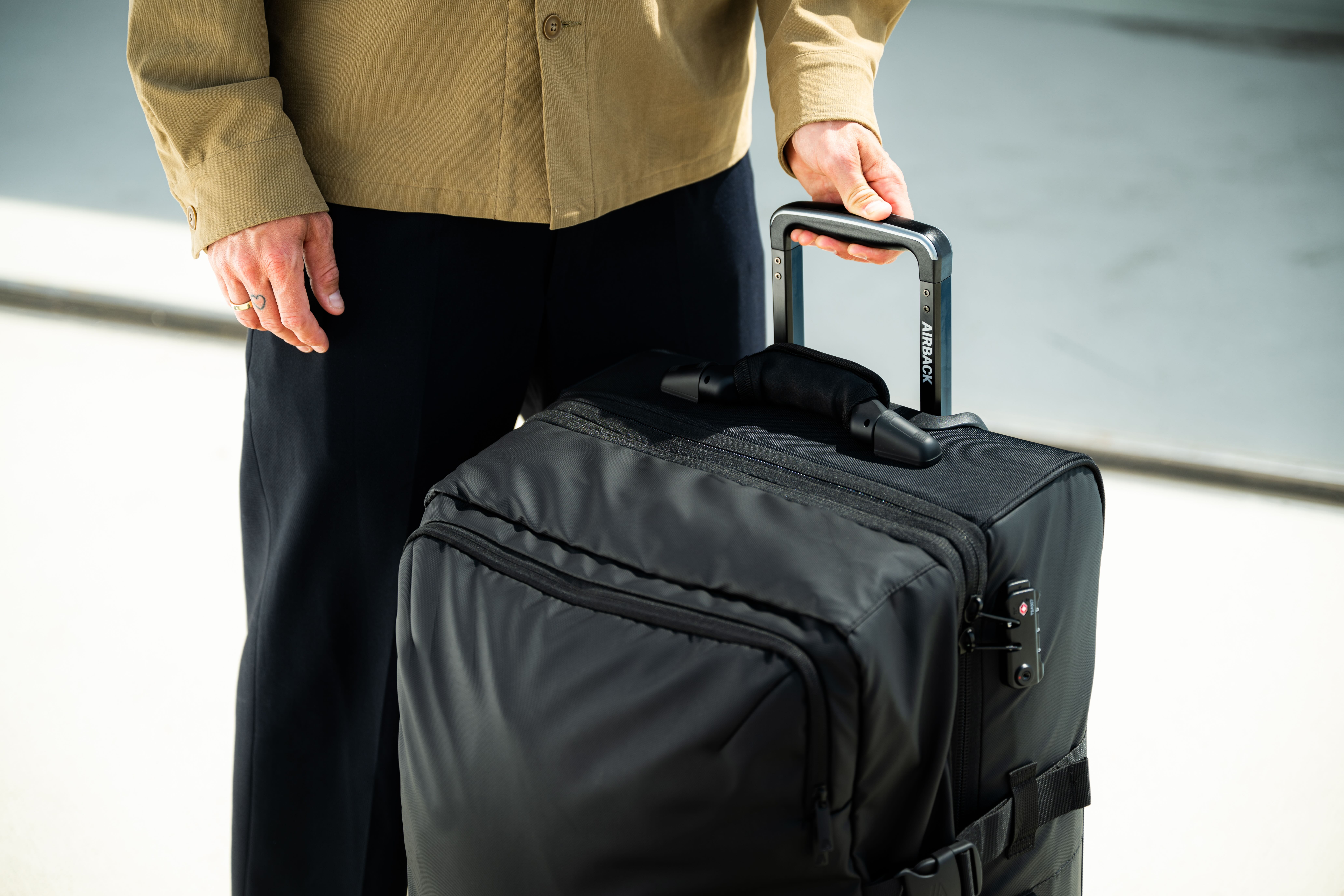 A person in a tan jacket holds the handle of a black rolling suitcase.