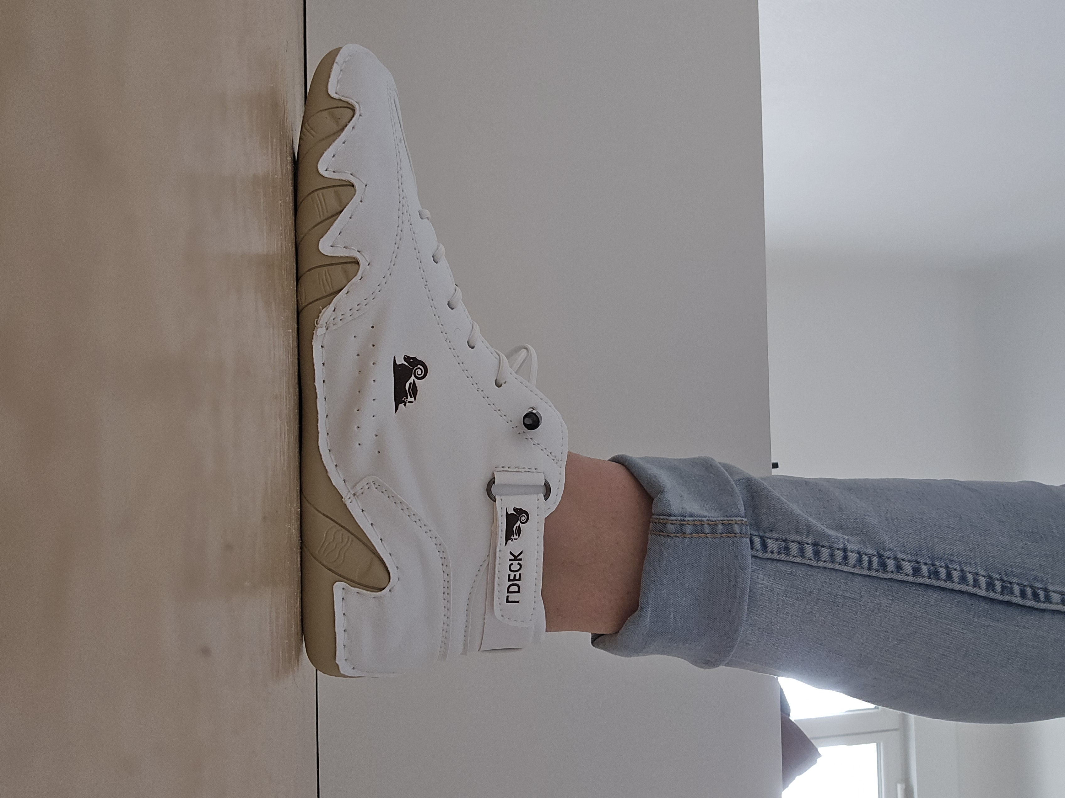 Person wearing white sneakers with a ram logo, standing on a wooden floor.