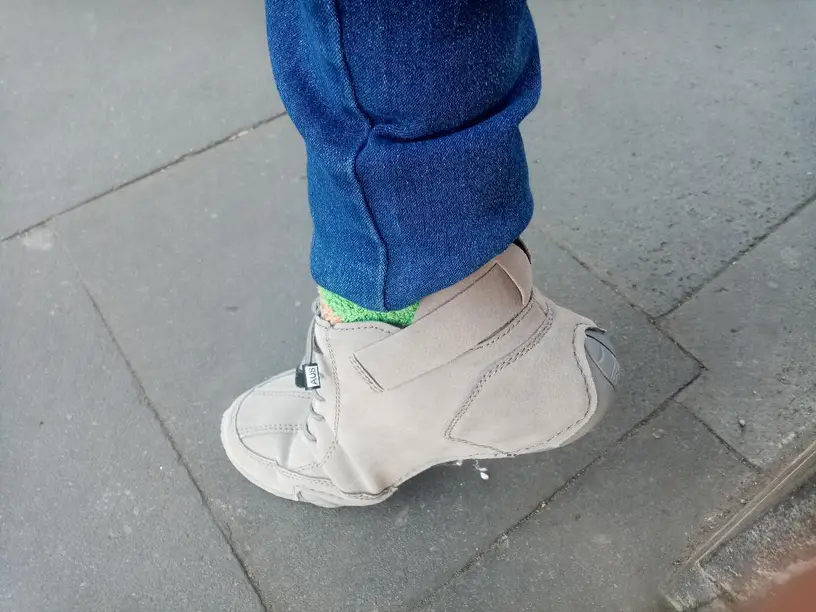 Person wearing blue jeans and beige orthopedic shoe on a sidewalk.