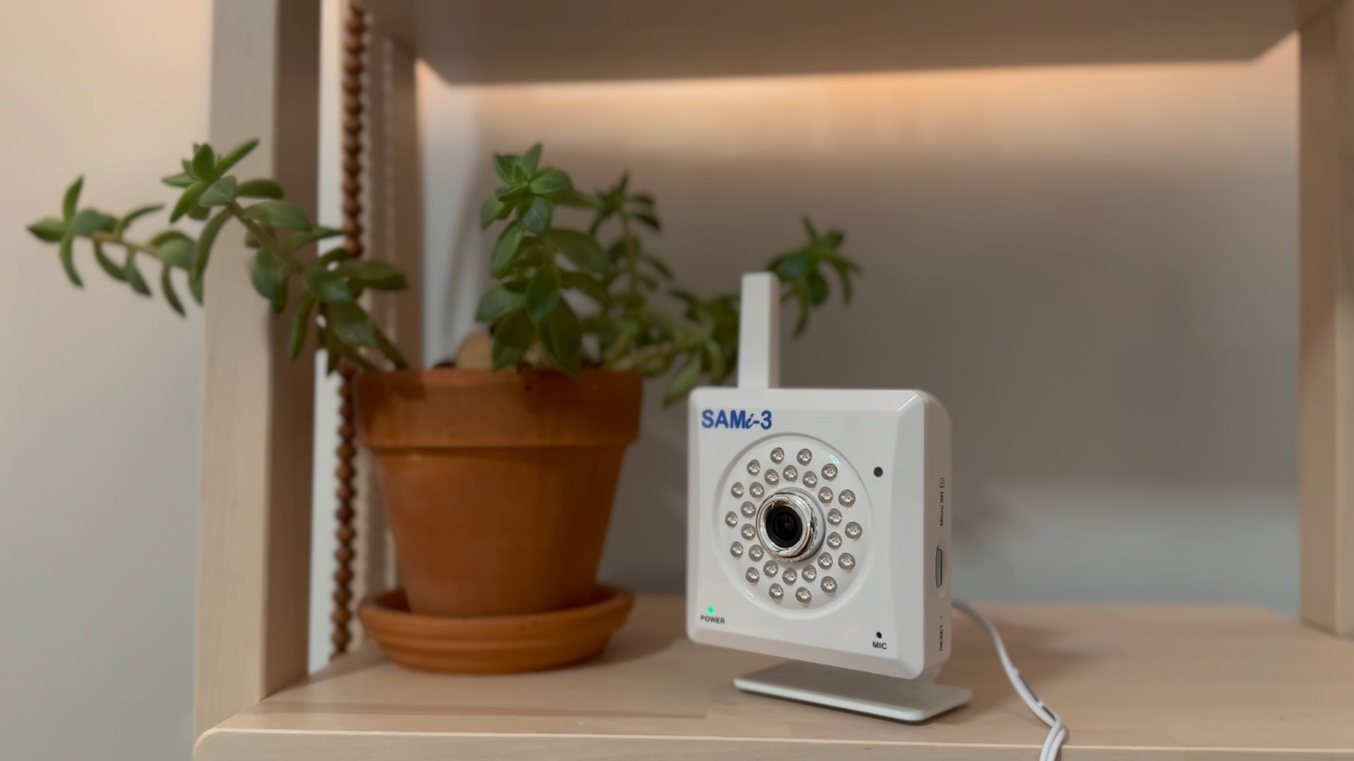 A white SAM-3 security camera sits on a wooden shelf next to a small potted plant.