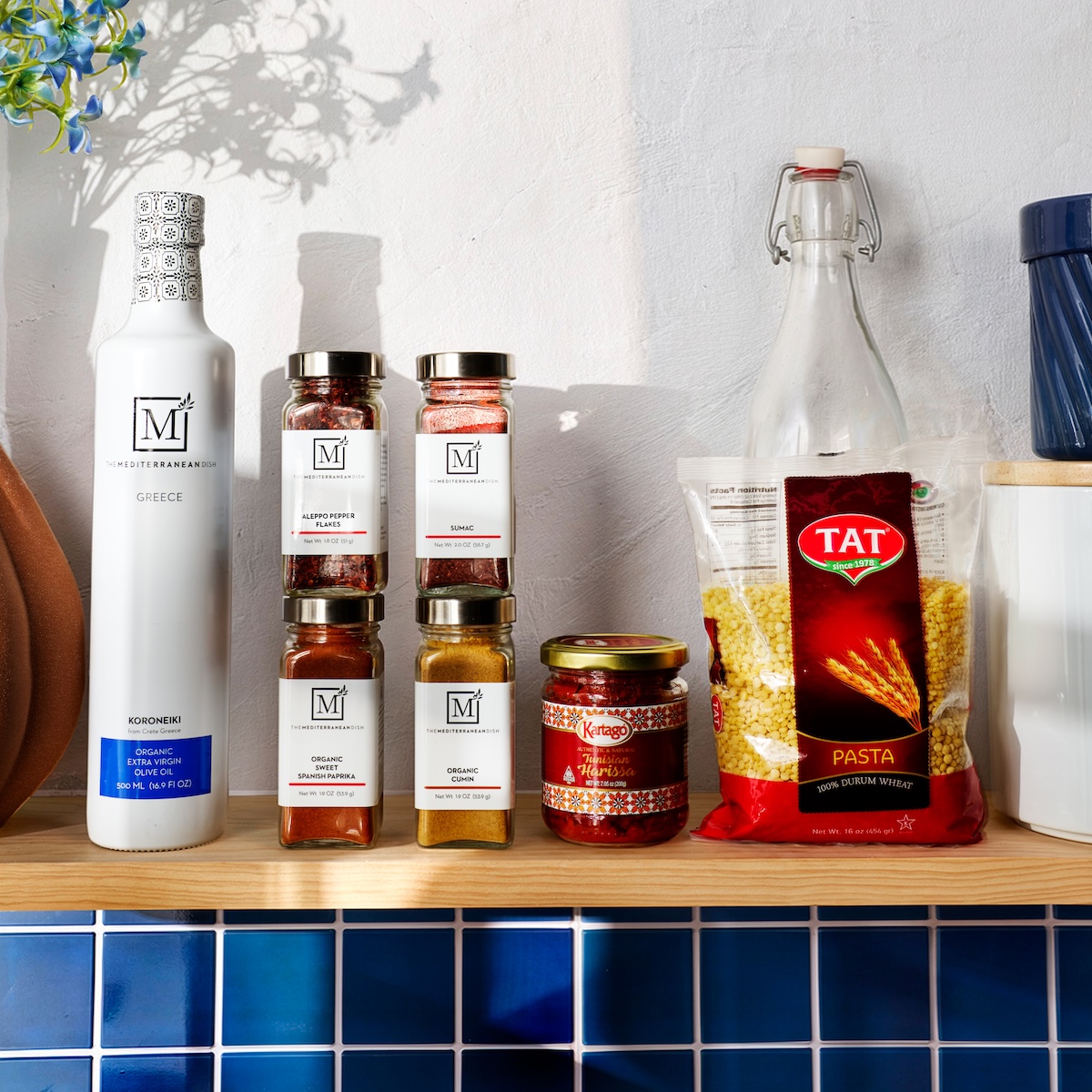 Olive oil, spices, harissa, and pasta arranged on a wooden shelf set against a blue tiled wall.