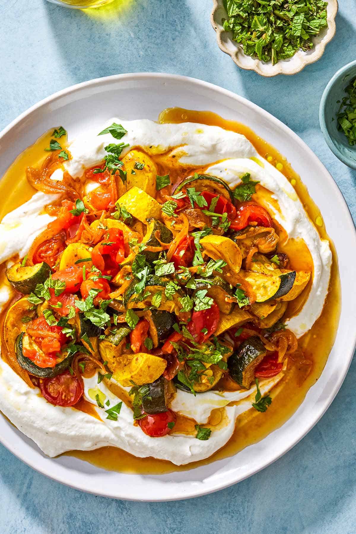 A white platter of sautéed squash, zucchini, and tomatoes served over a bed of creamy white cheese or yogurt.