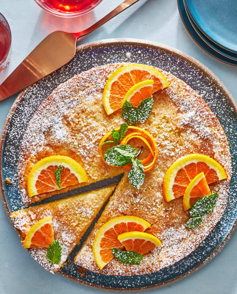 An overhead shot of a sliced orange cake, dusted with powdered sugar and garnished with mint leaves.