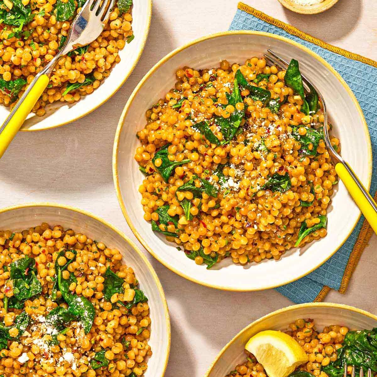 An overhead view of several bowls of pearl couscous with spinach and grated cheese.