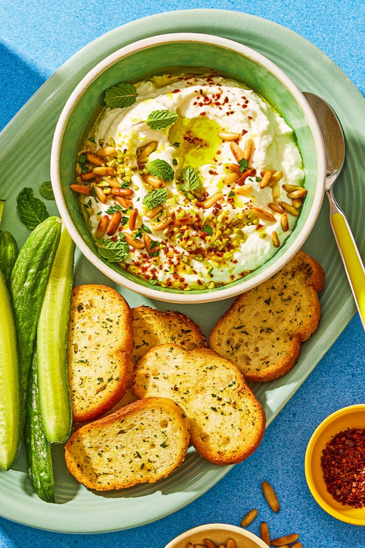 An overhead shot of a creamy dip with pine nuts and mint, served with toasted bread and cucumbers.