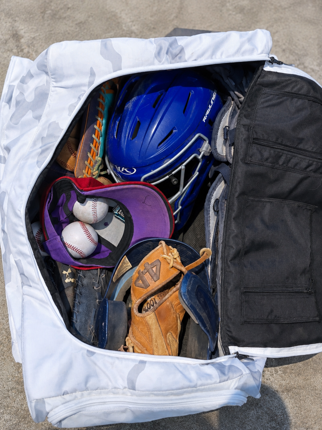 An open white camouflage duffel bag filled with baseball equipment, including a helmet, gloves, and baseballs.