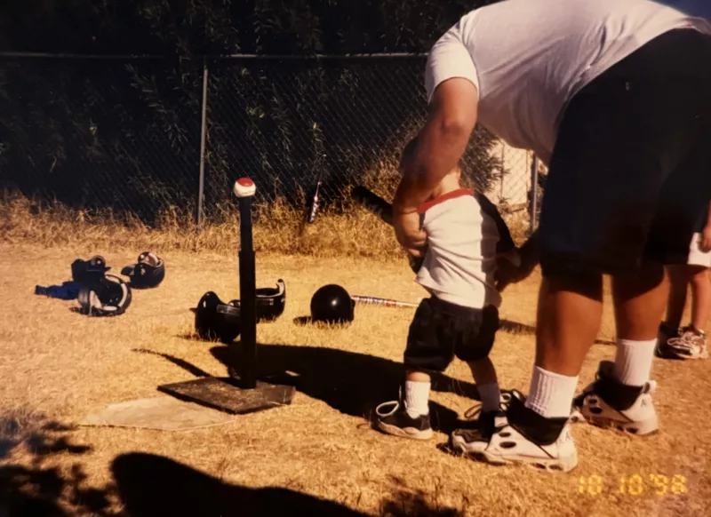 An adult helps a young child swing a baseball bat at a ball on a tee.