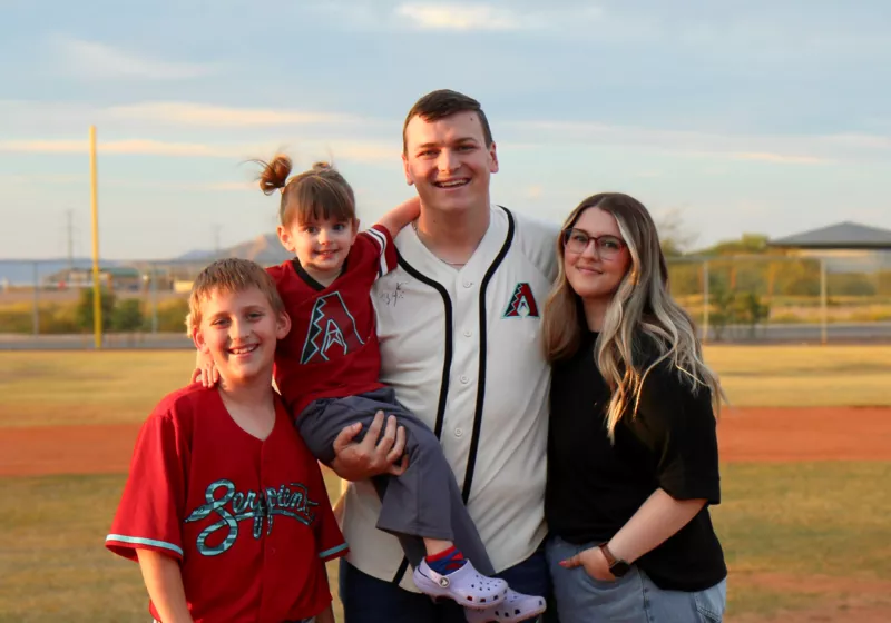 A family of four, two adults and two children, pose for a photo on a baseball field.