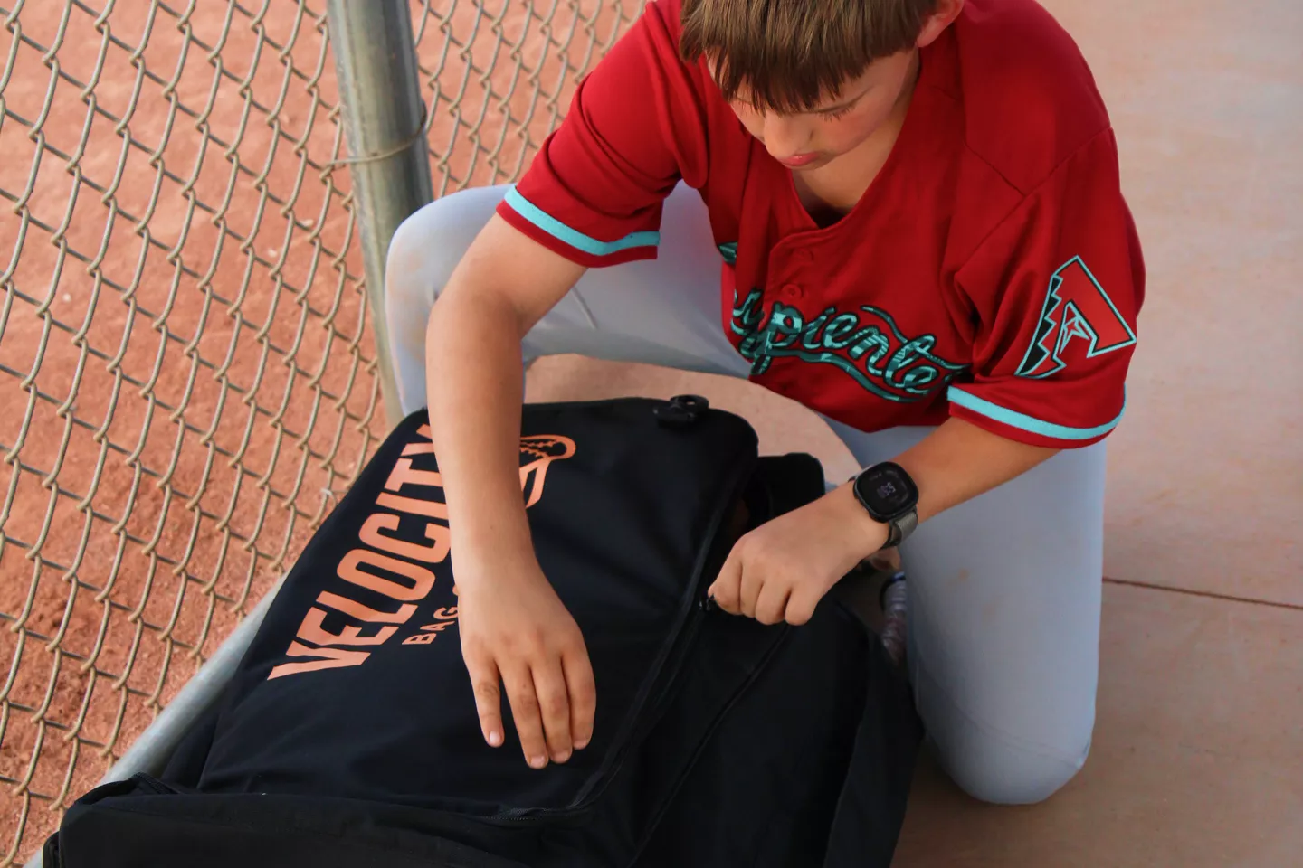A young baseball player in a red jersey kneels and zips open a large black Velocity sports bag.