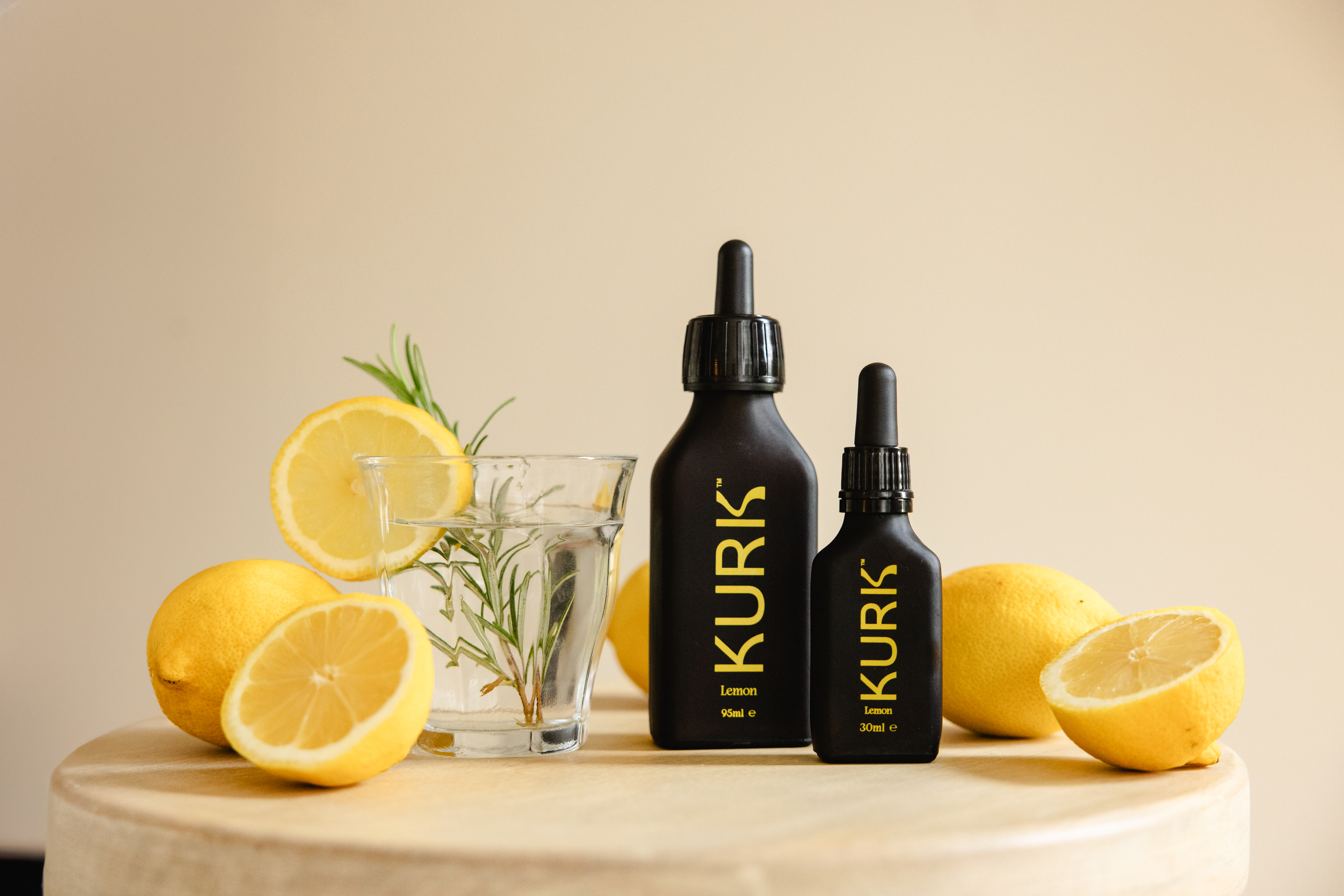 Two black KURK lemon dropper bottles next to a glass of water with rosemary and fresh lemons.
