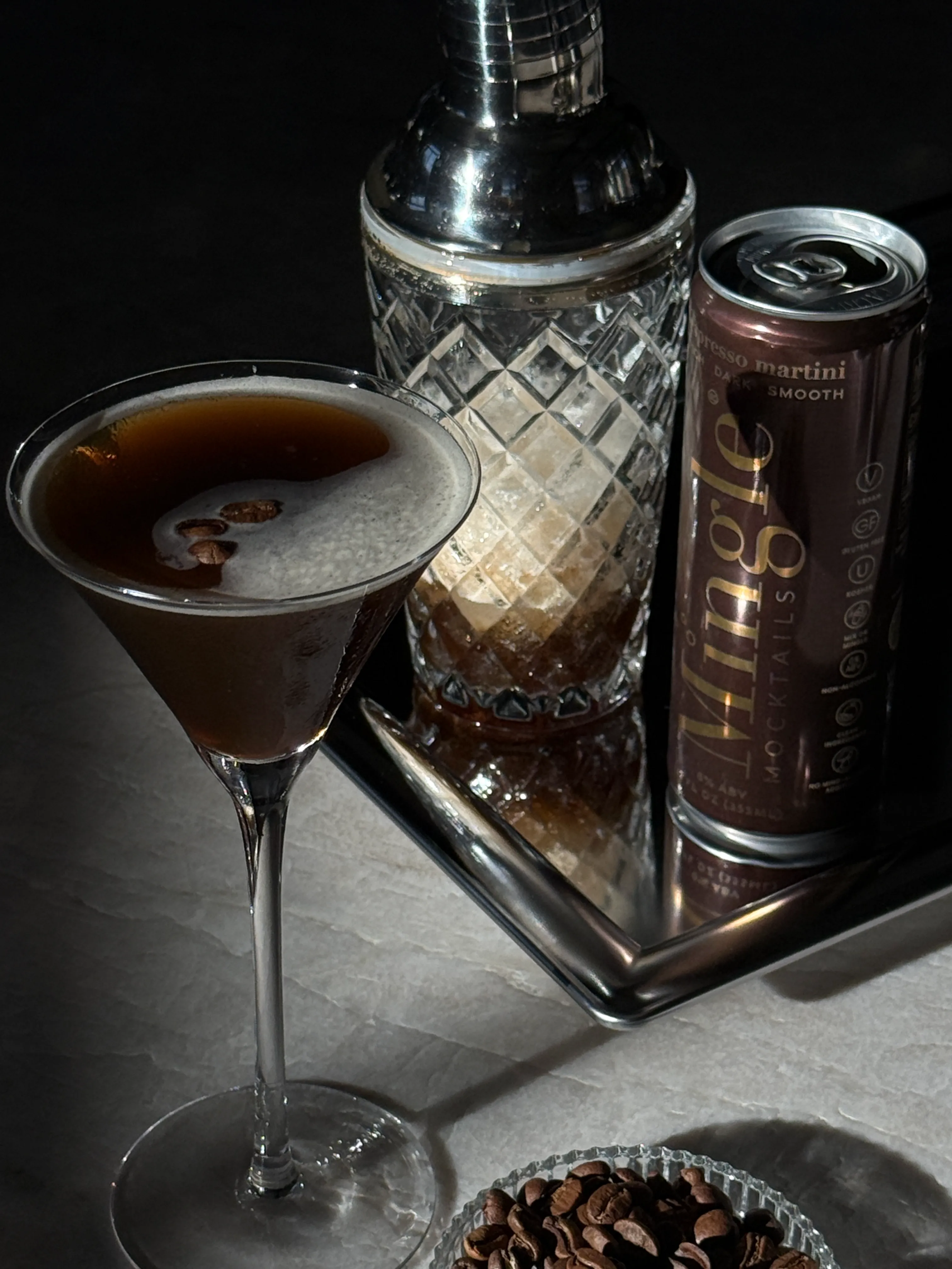 An espresso martini mocktail in a glass on a silver tray with a shaker and a can.
