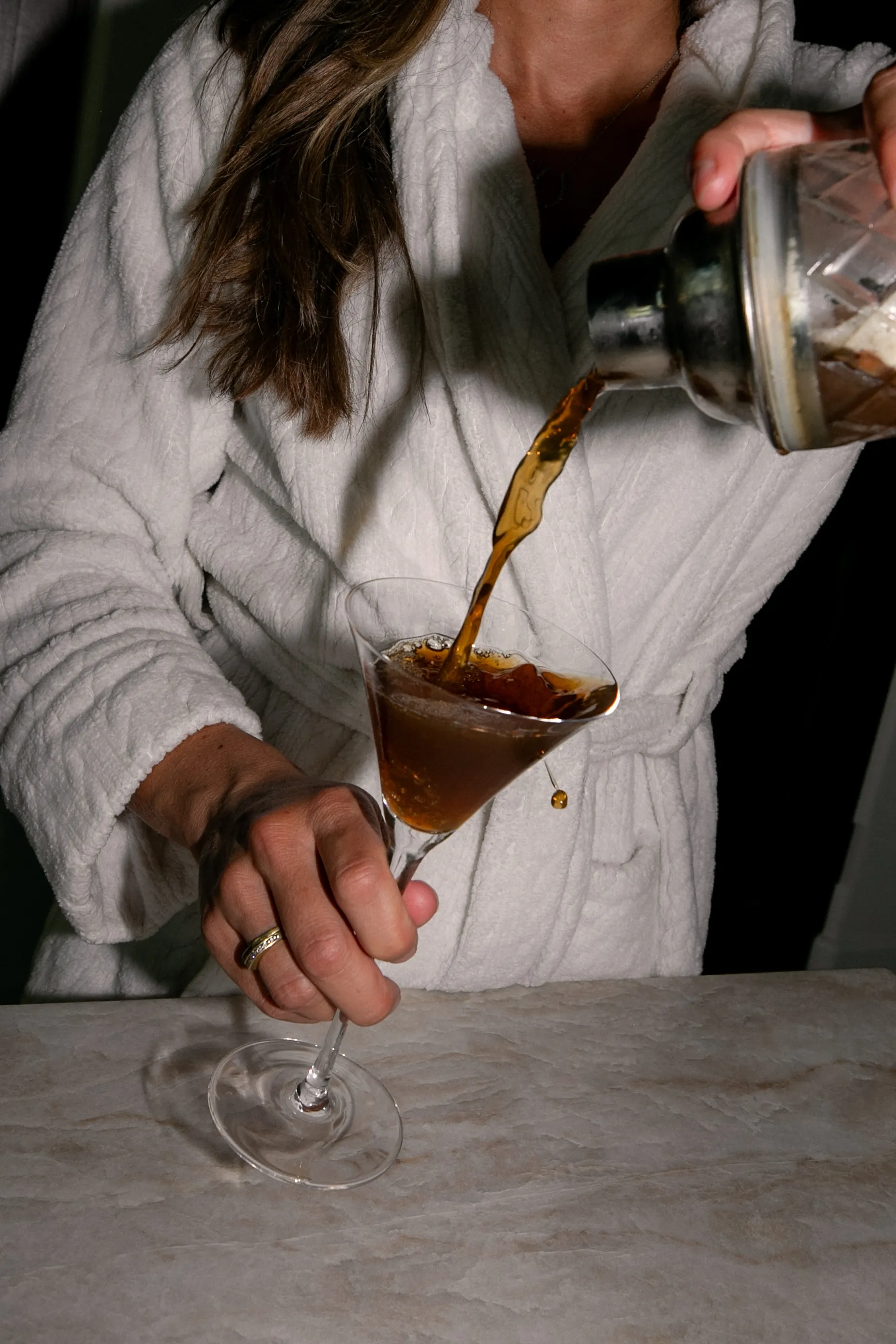 A person in a white bathrobe pours a dark drink from a shaker into a martini glass.