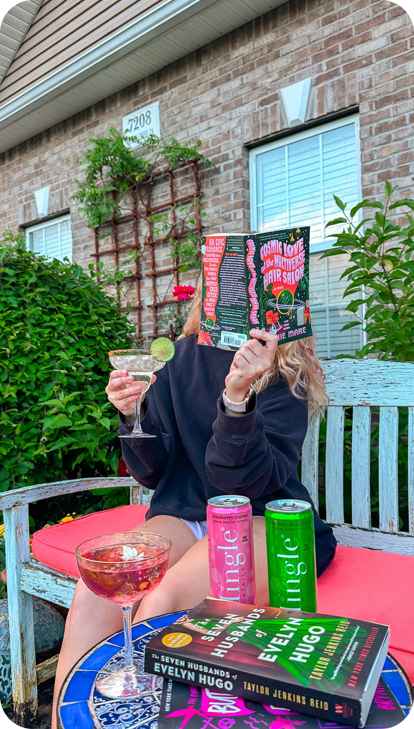 A person sits on a bench outside, holding a cocktail and hiding their face with a book.