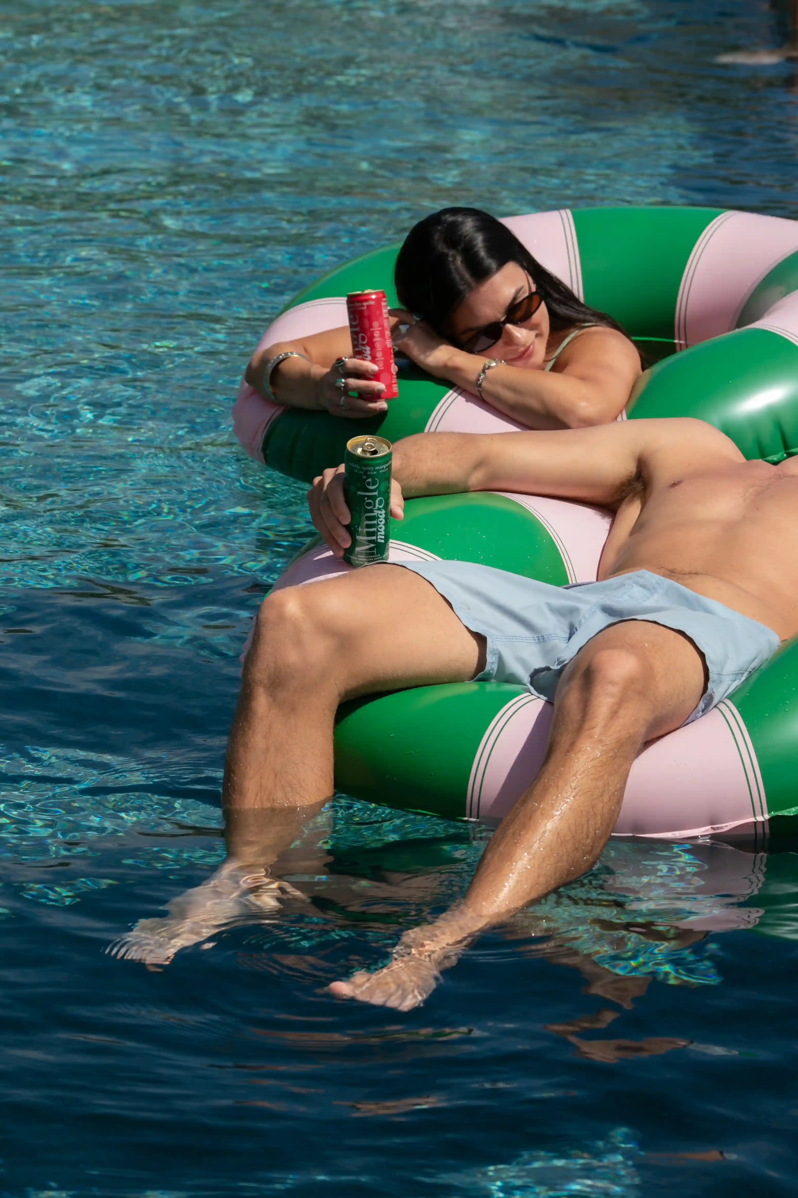 A man and a woman relax on a large inflatable ring in a swimming pool, holding cans.