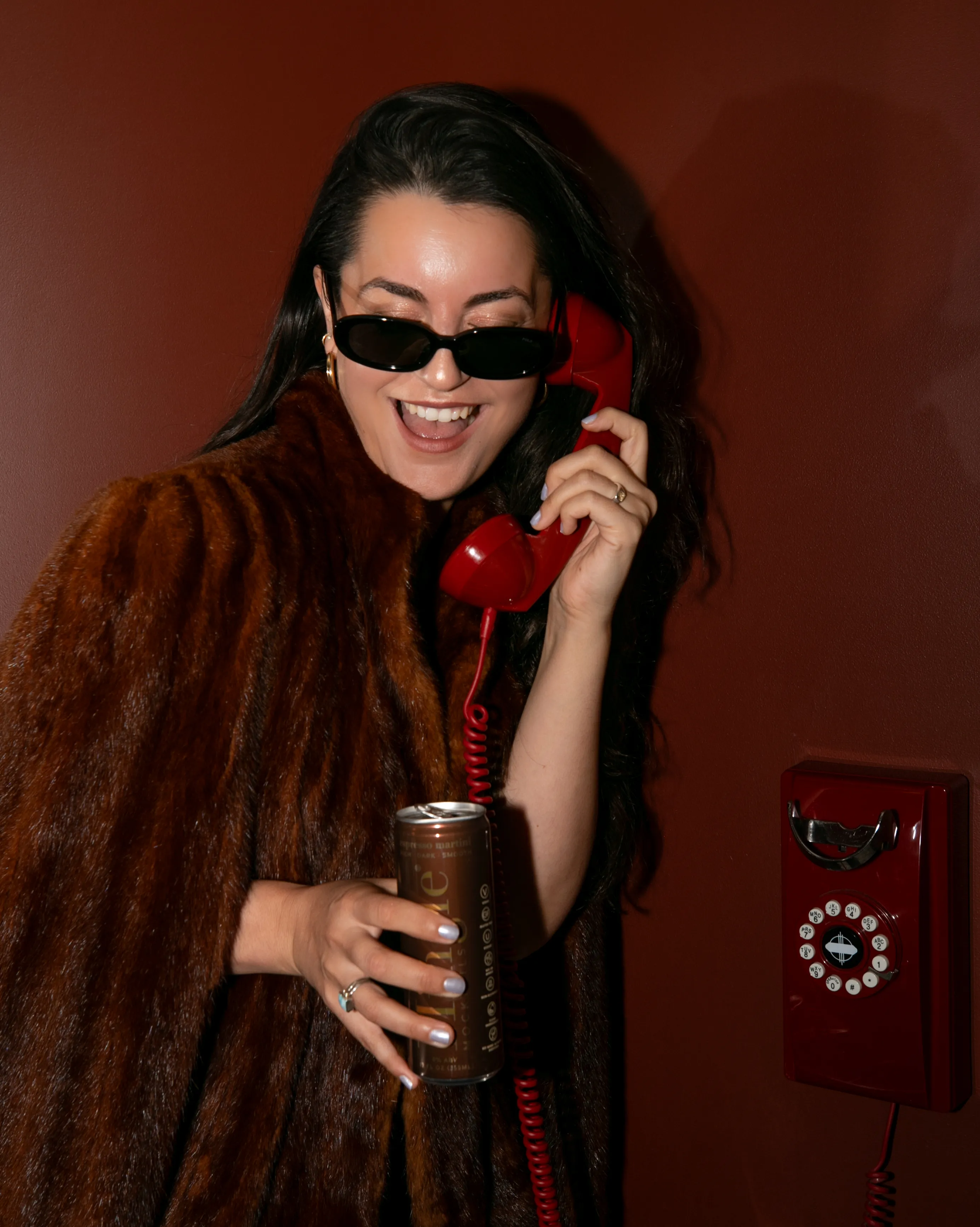 A woman in a fur coat and sunglasses smiles while on a red rotary phone, holding a can.