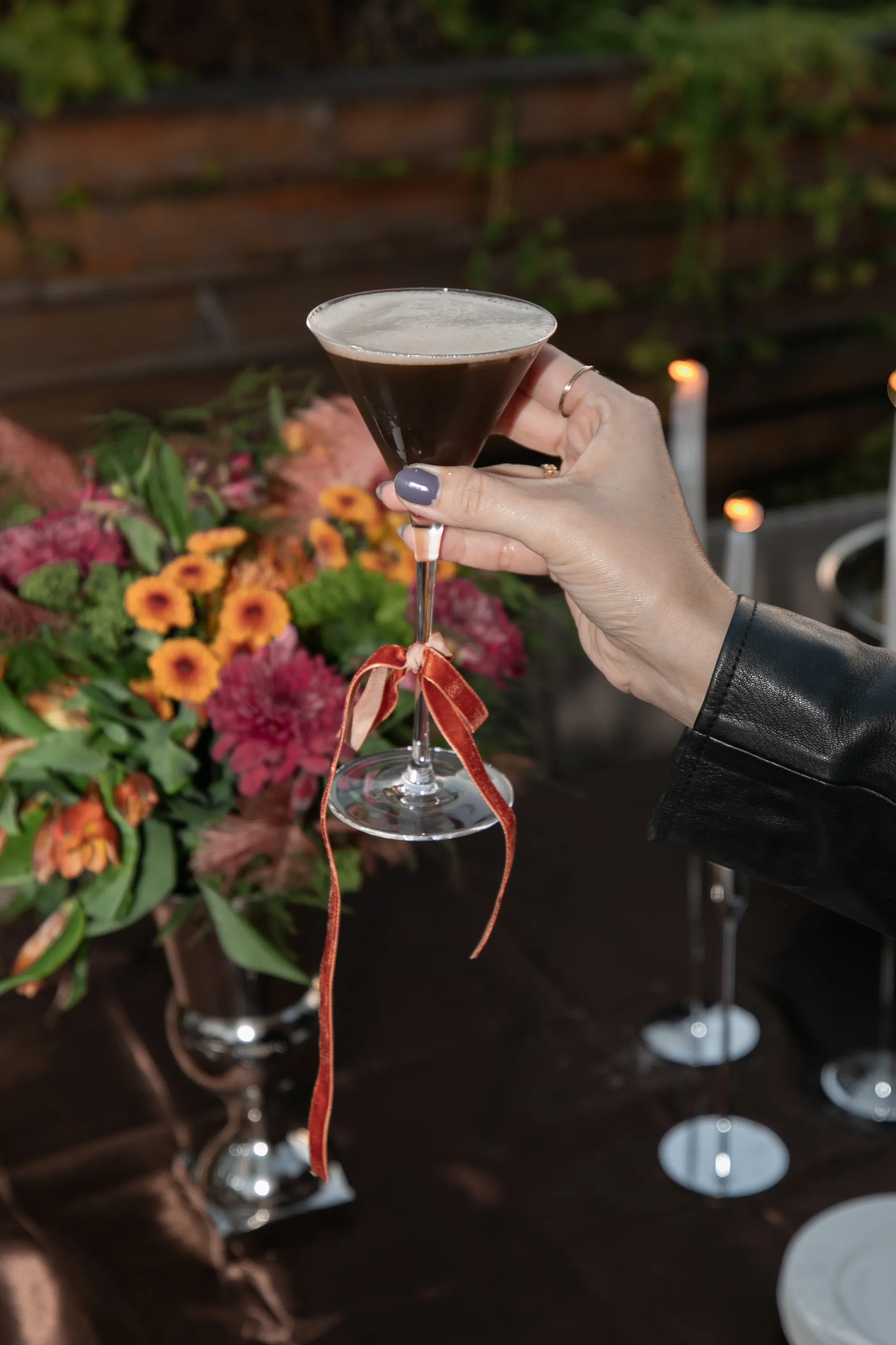 A hand holds a dark, foamy cocktail in a martini glass with a red ribbon on the stem.